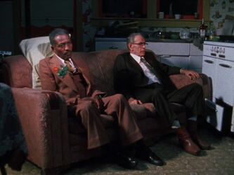 Leviticus "Rose" Washington and Ernie Neuman from John Huckert's The Passing (1983) sitting in funeral attire on a couch inexplicably moved to their kitchen.