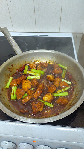 A chicken dish, sizzling away in a stainless steel pan. It's cooked in a deep brown, savoury sauce with vibrant green spring onion and dried chilli.