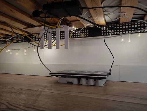 A picture taken underneath a bed, with a laptop ontop of some egg cartons on the floor. There's two cables going up towards the bed, that have a router/ap combination tied onto the bottom of the bed. There's some fairy lights in theback providing the light for the picture.
