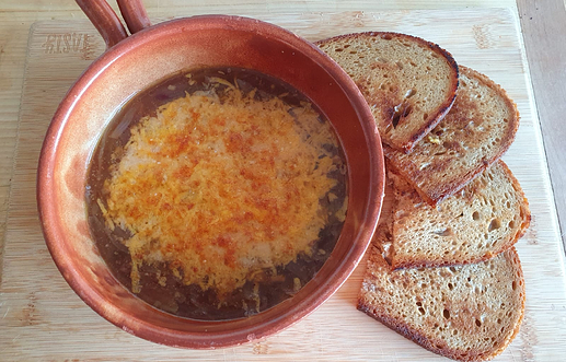 A bowl with a brown soup with molted cheese on top. On the left next to the bowl are four slightly roasted slices of bread.
