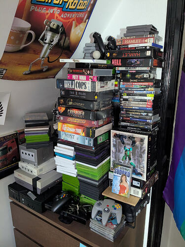 Poorly balanced stacks of DVD cases and big box PC games, along with several game consoles, cartridges, and controllers.