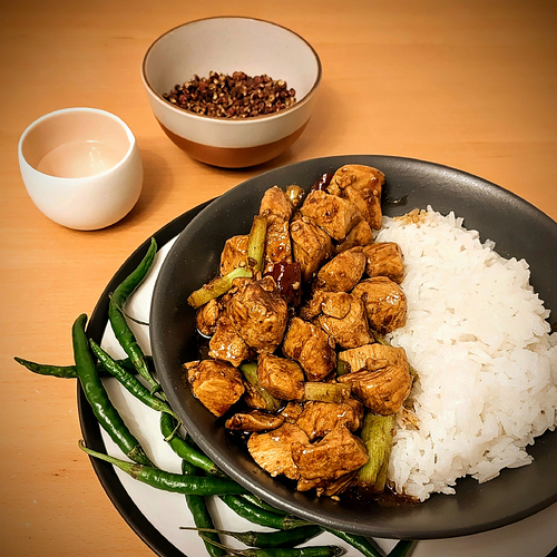 Photograph of fried chicken in a deep brown sauce with spring onion and dried chillies, served with jasmine rice. It's set within a grey bowl atop a white plate with black rim. In the background a small pot of water and bowl of Sichuan Pepper are visible. It rests on a wooden table