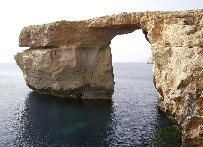a gigantic sea arch. The sea is calm, and the sky clear and white.