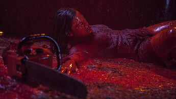 Screenshot from Evil Dead 2013. Mia lies on the floor, soaked in blood whilst blood rain falls. Her arm is outstretched towards a chainsaw