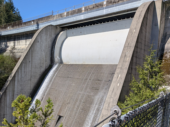 Picture of a large concrete dam's outlet. At the centre there's a white round metal gate that's closed with only small amounts of water leaking through. There's a walkway above it following the curve and some greenery surrounding the imaged structure.