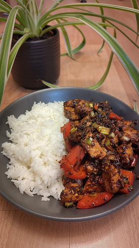 Photo of a chicken-based dish with pepper and spring onion stir fried with fermented soybean paste, dark soy, ginger, garlic, sichuan pepper, dried chillies, sugar and salt. It's served in a large grey bowl with a generous portion of jasmine rice