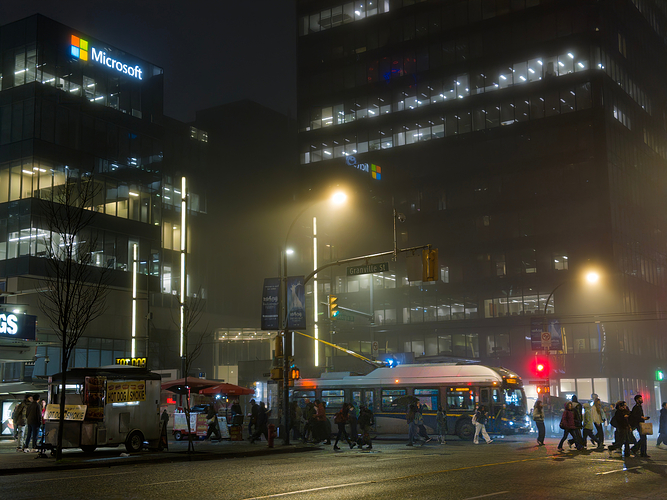 A downtown Vancouver intersection on a foggy evening. Numerous pedestrians and a trolleybus can be seen crossing the street against the backdrop of skyscrapers and hot dog stands.