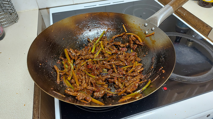 an enormous wok, filled with a beef and celery stir fry. It sits atop a cheap, ill-fitted stovetop with visible gaps and unfinished plywood edges on the surrounding counter